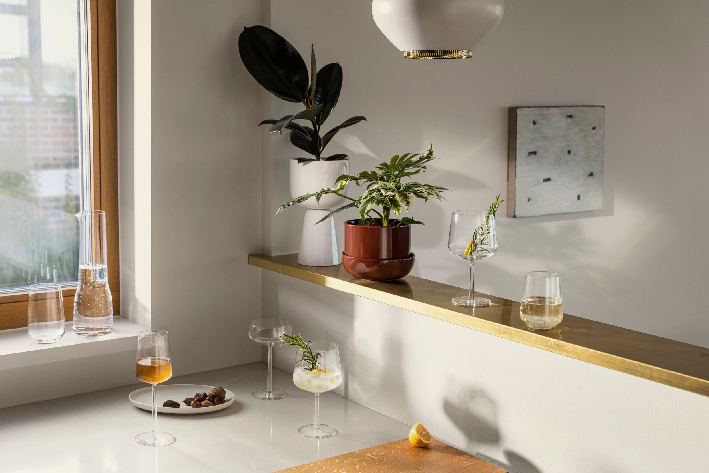 Ett modernt kök med cocktails och vin på vita bänkskivor och en gyllene hylla, prydd med krukväxter och minimalistisk konst.