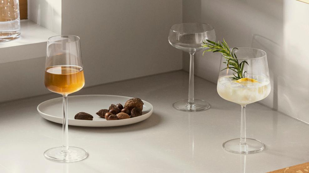 Ett modernt kök med cocktails och vin på vita bänkskivor och en gyllene hylla, prydd med krukväxter och minimalistisk konst.