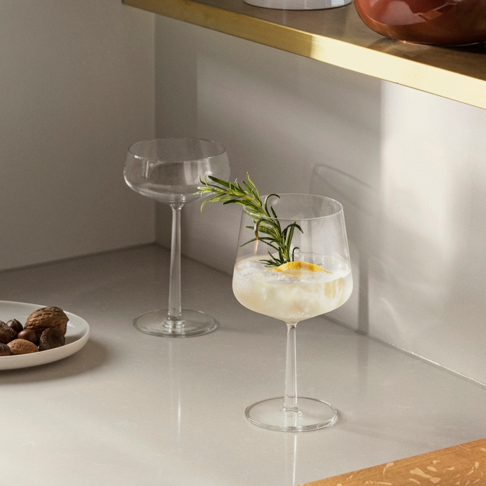 Ett modernt kök med cocktails och vin på vita bänkskivor och en gyllene hylla, prydd med krukväxter och minimalistisk konst.