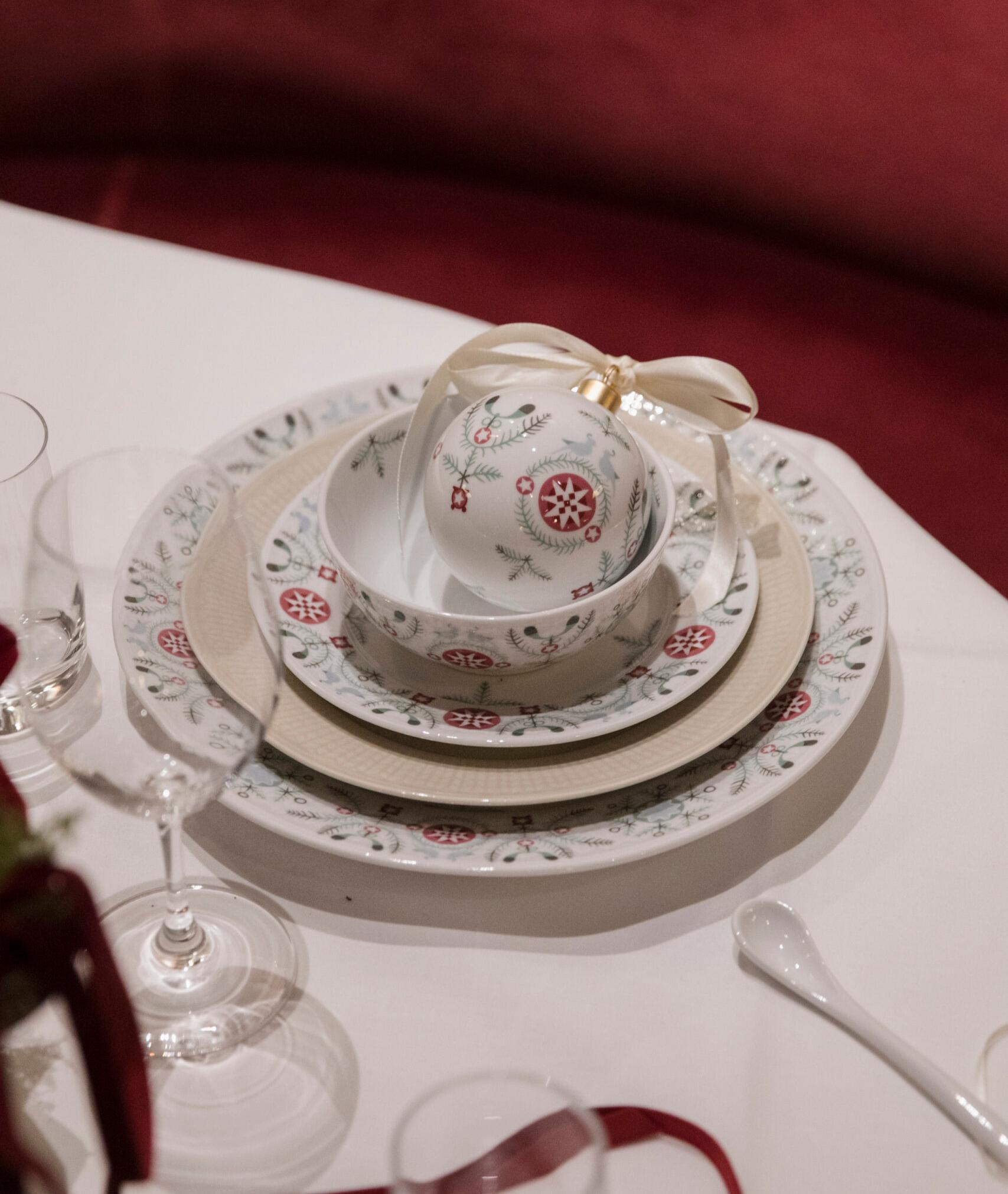 Juhlava kattaus kuviollisilla lautasilla, kulholla ja koristeellisella joulukoristeella, jossa on nauha.