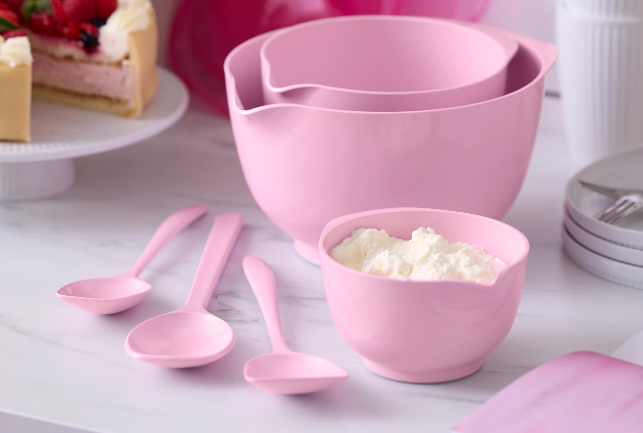 Rosa bakredskap med skålar och slevar står på en köksbänk bredvid en gräddtårta med färska bär.