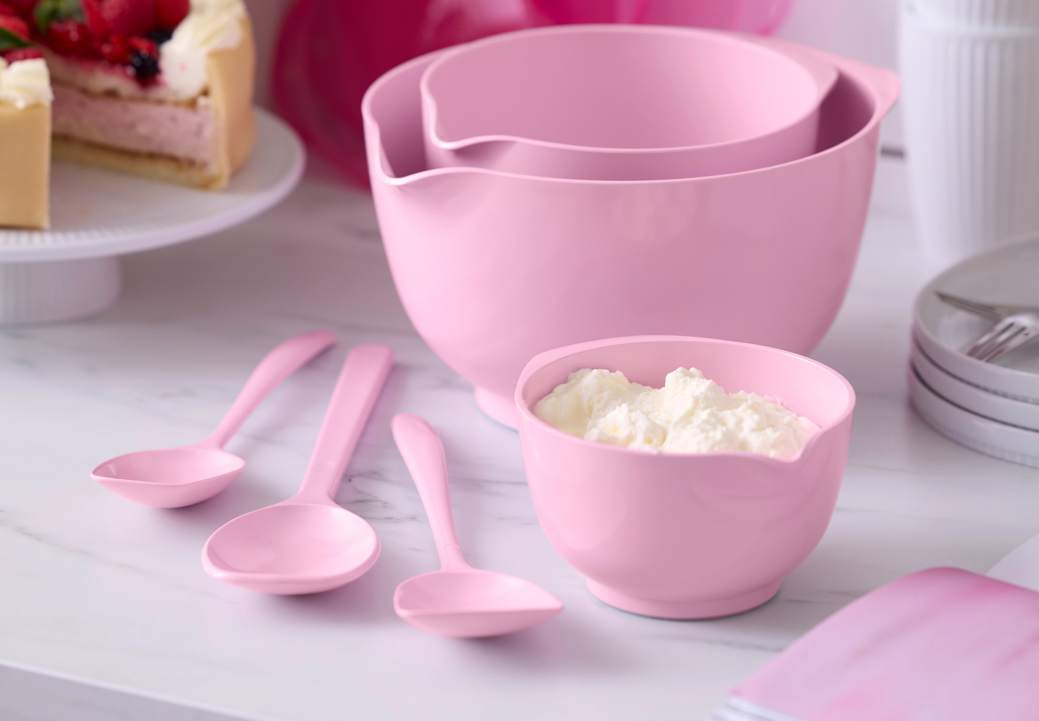 Rosa bakredskap med skålar och slevar står på en köksbänk bredvid en gräddtårta med färska bär.