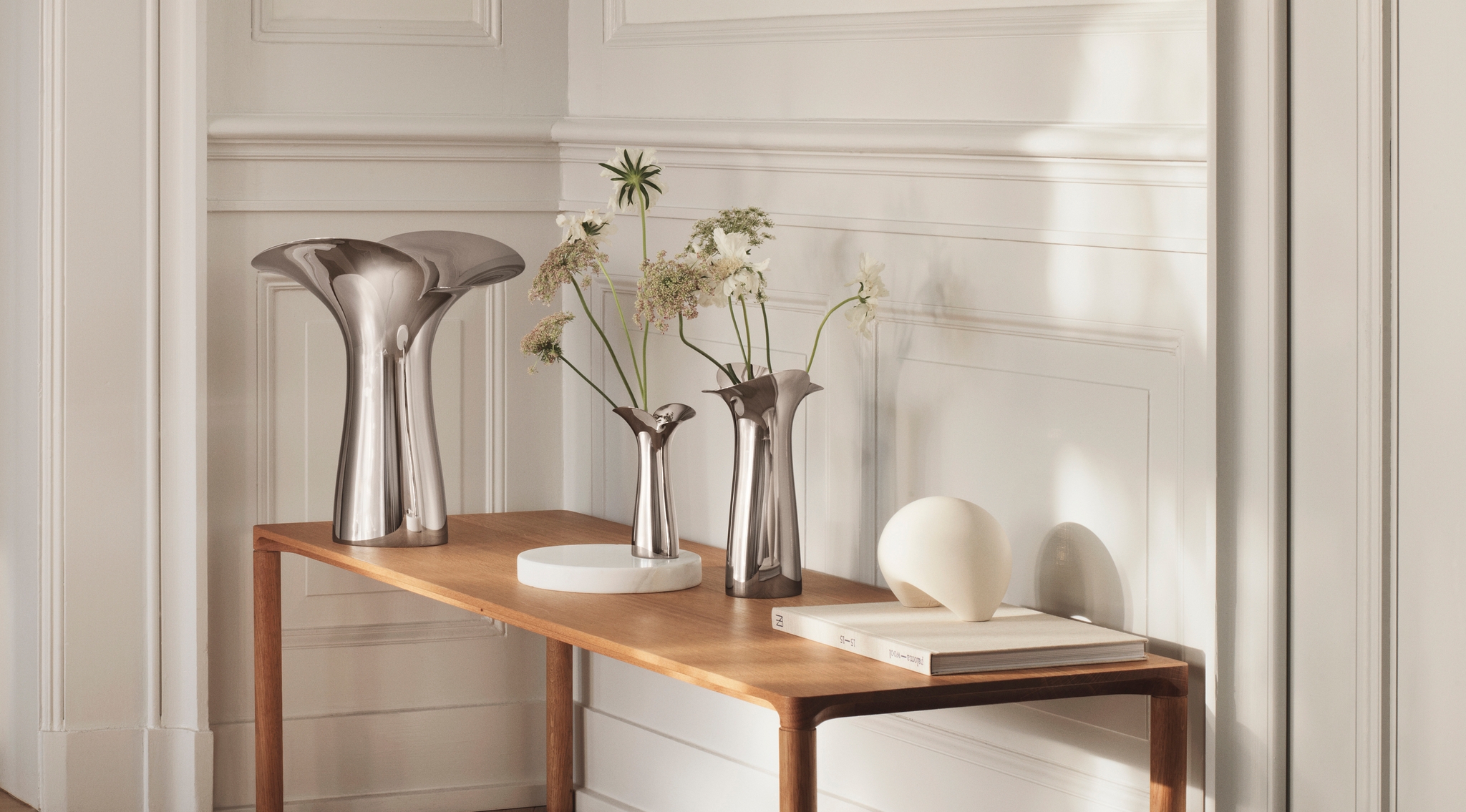Lyst, minimalistisk interiør med en trebenk mot en hvit vegg, dekorert med moderne metallvaser og blomster.