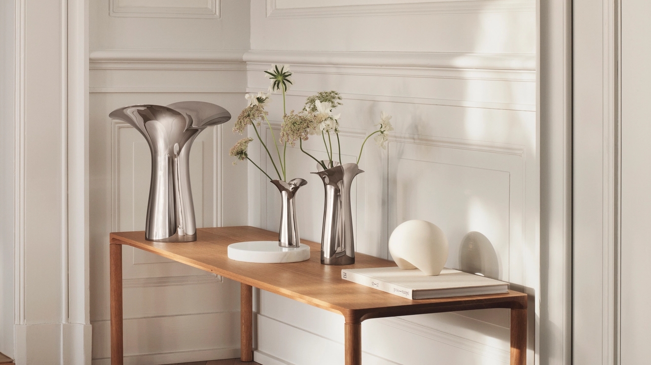 Ljus, minimalistisk hall med träbänk, skulpturala metallvaser och torkade blommor mot vit panelvägg.