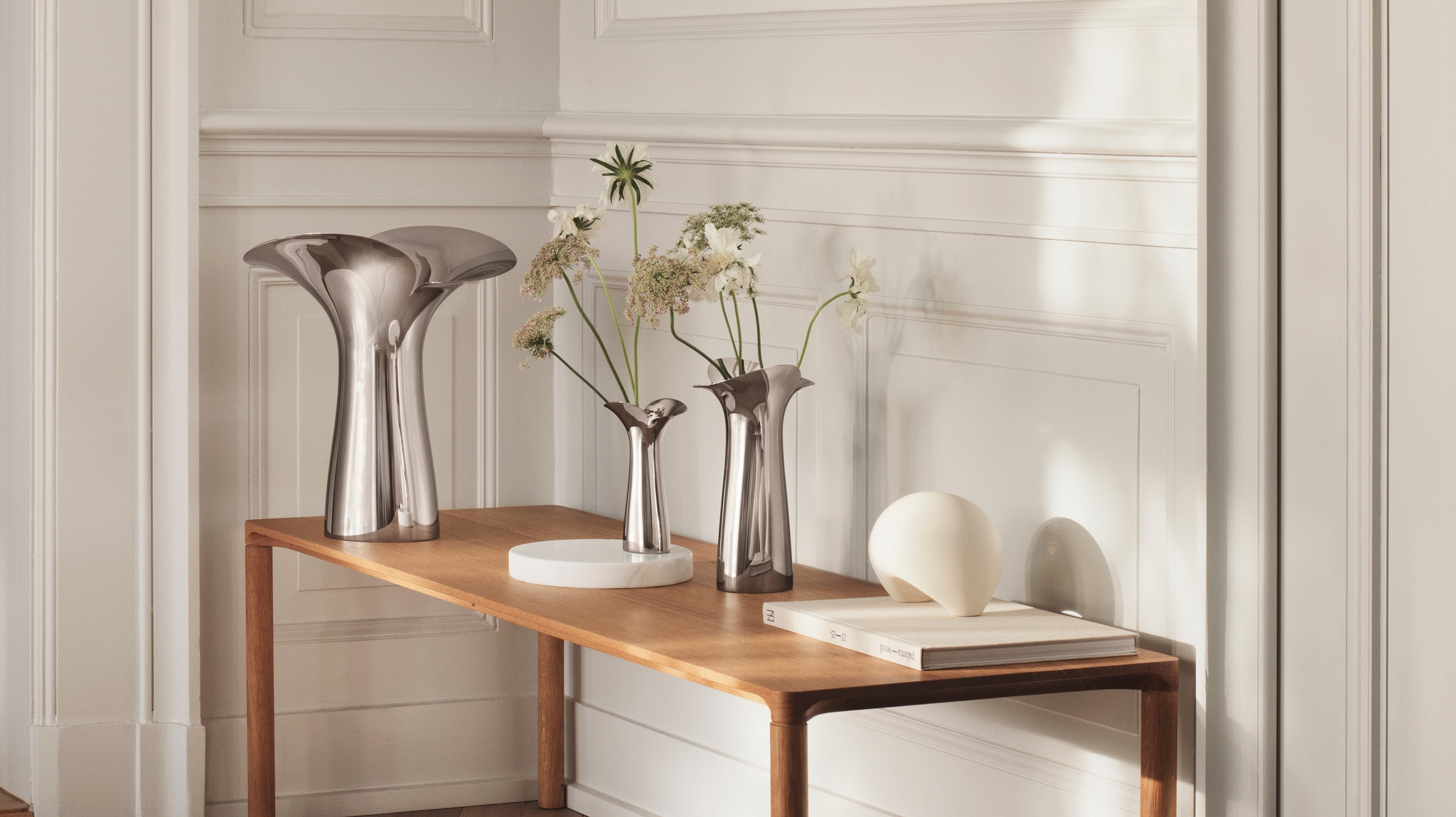 Ljus, minimalistisk hall med träbänk, skulpturala metallvaser och torkade blommor mot vit panelvägg.