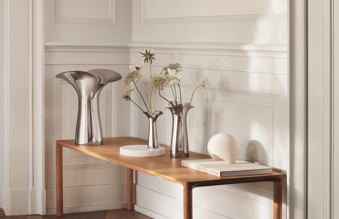 Ljus, minimalistisk hall med träbänk, skulpturala metallvaser och torkade blommor mot vit panelvägg.