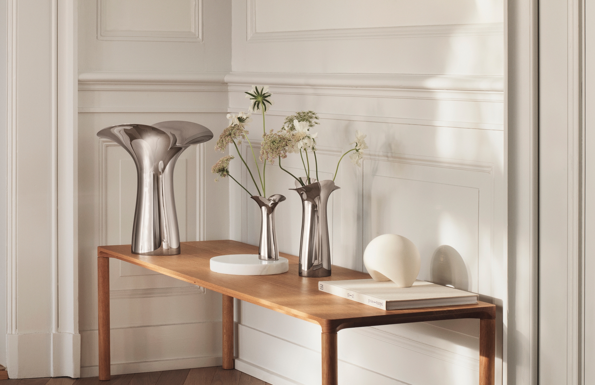 Ljus, minimalistisk hall med träbänk, skulpturala metallvaser och torkade blommor mot vit panelvägg.