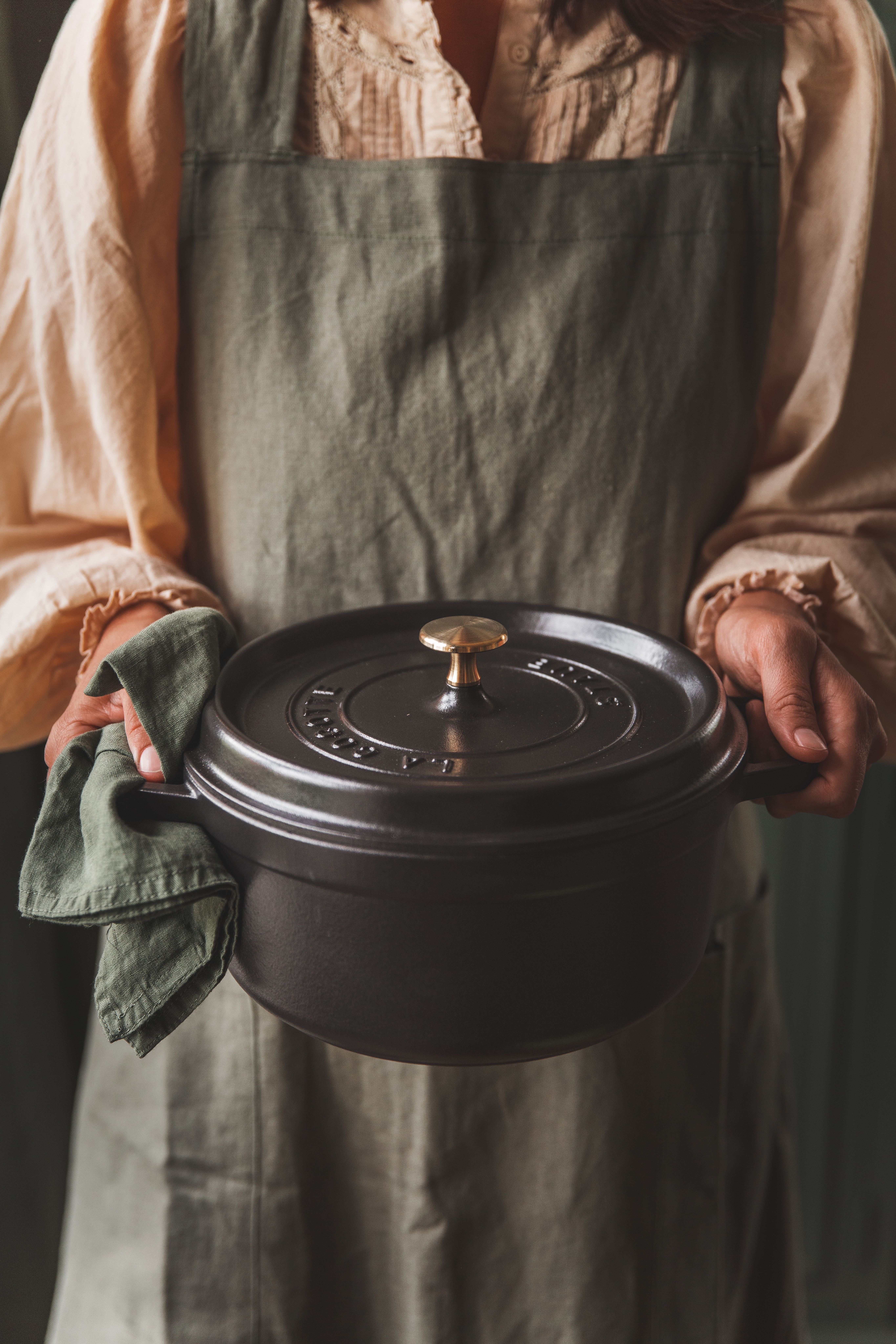Esiliinapäinen henkilö pitelee tummaa valurautaista pataa ja vihreää pellavapyyhettä.