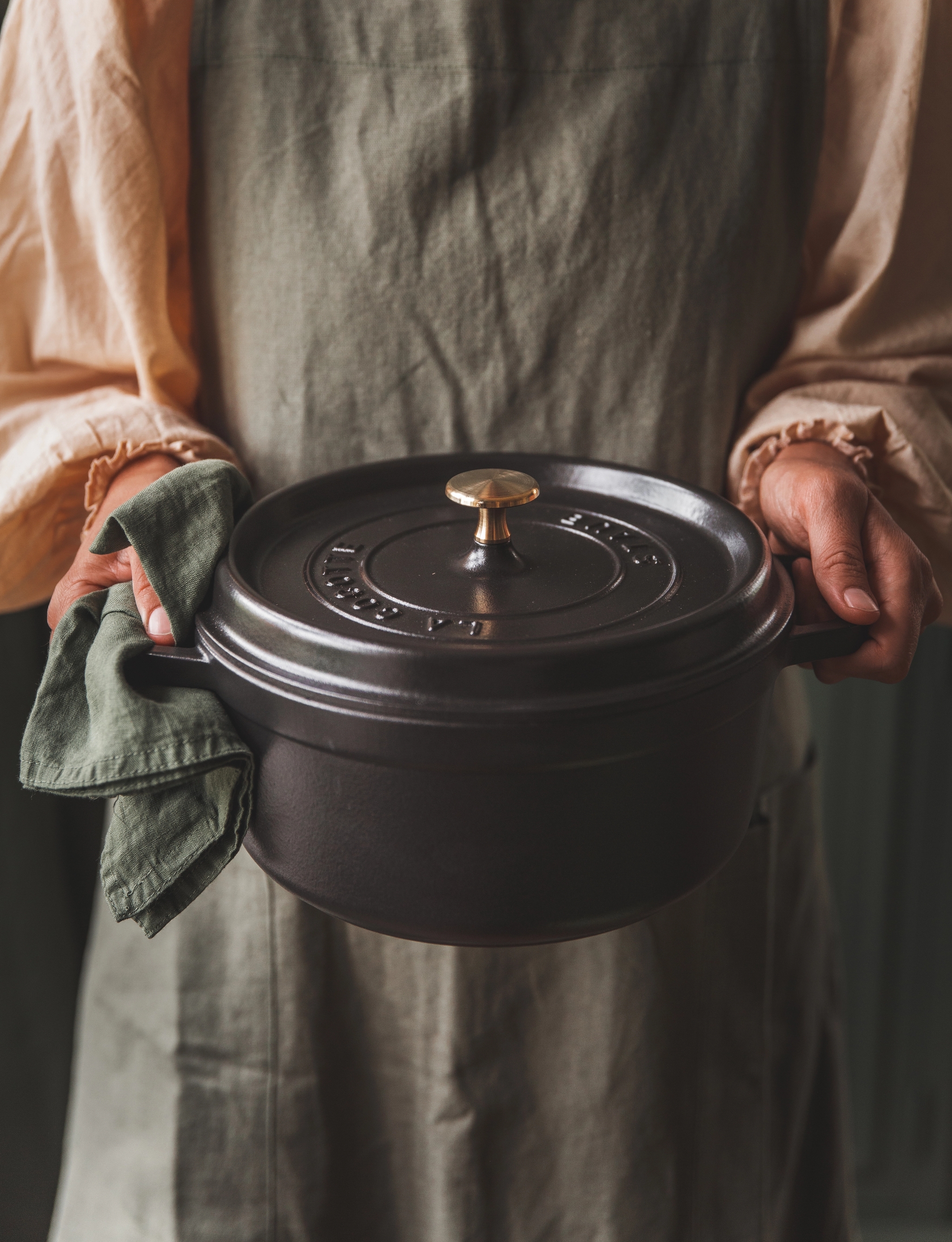 Esiliinapäinen henkilö pitelee tummaa valurautaista pataa ja vihreää pellavapyyhettä.
