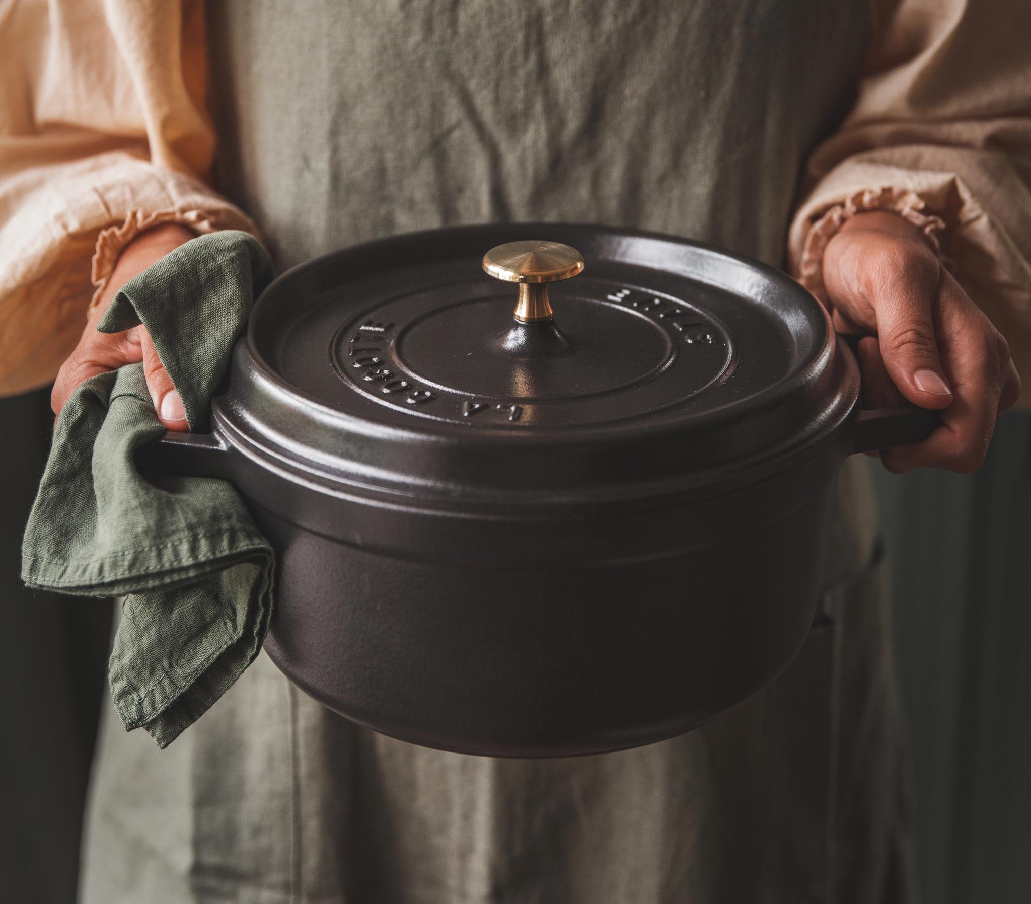Esiliinapäinen henkilö pitelee tummaa valurautaista pataa ja vihreää pellavapyyhettä.