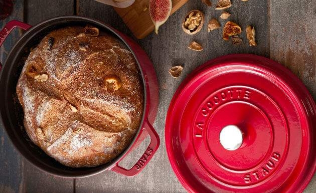 Punaisessa valurautapadassa paistettu tuore leipä on katettuna pöydälle yhdessä juustojen, viikunoiden ja saksanpähkinöiden kanssa.
