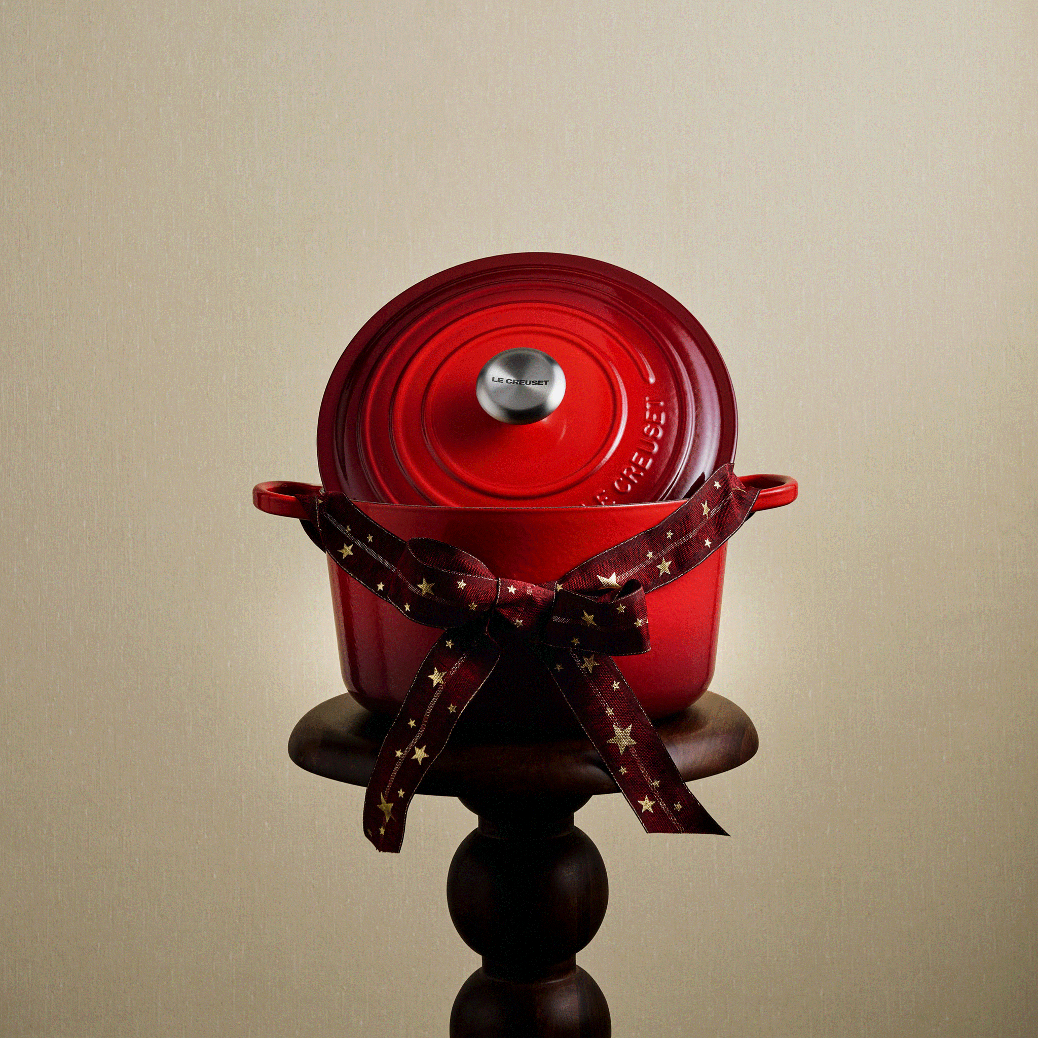 Red Le Creuset Dutch oven with lid, tied with a star-patterned ribbon, on a wooden stand.