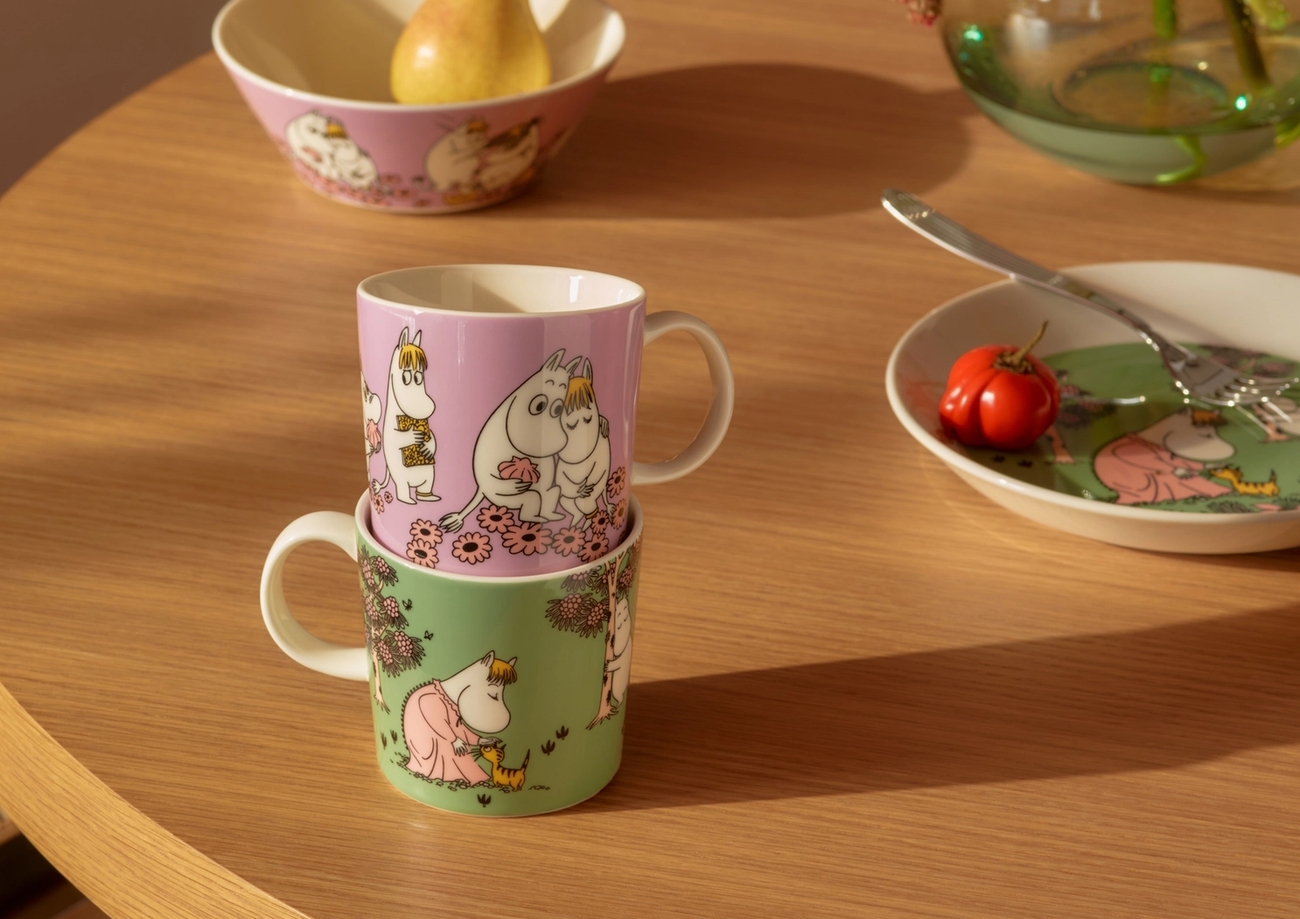 Kaksi päällekkäin asetettua Muumimukia ja Muumikulho, jossa on päärynä, puisella pöydällä