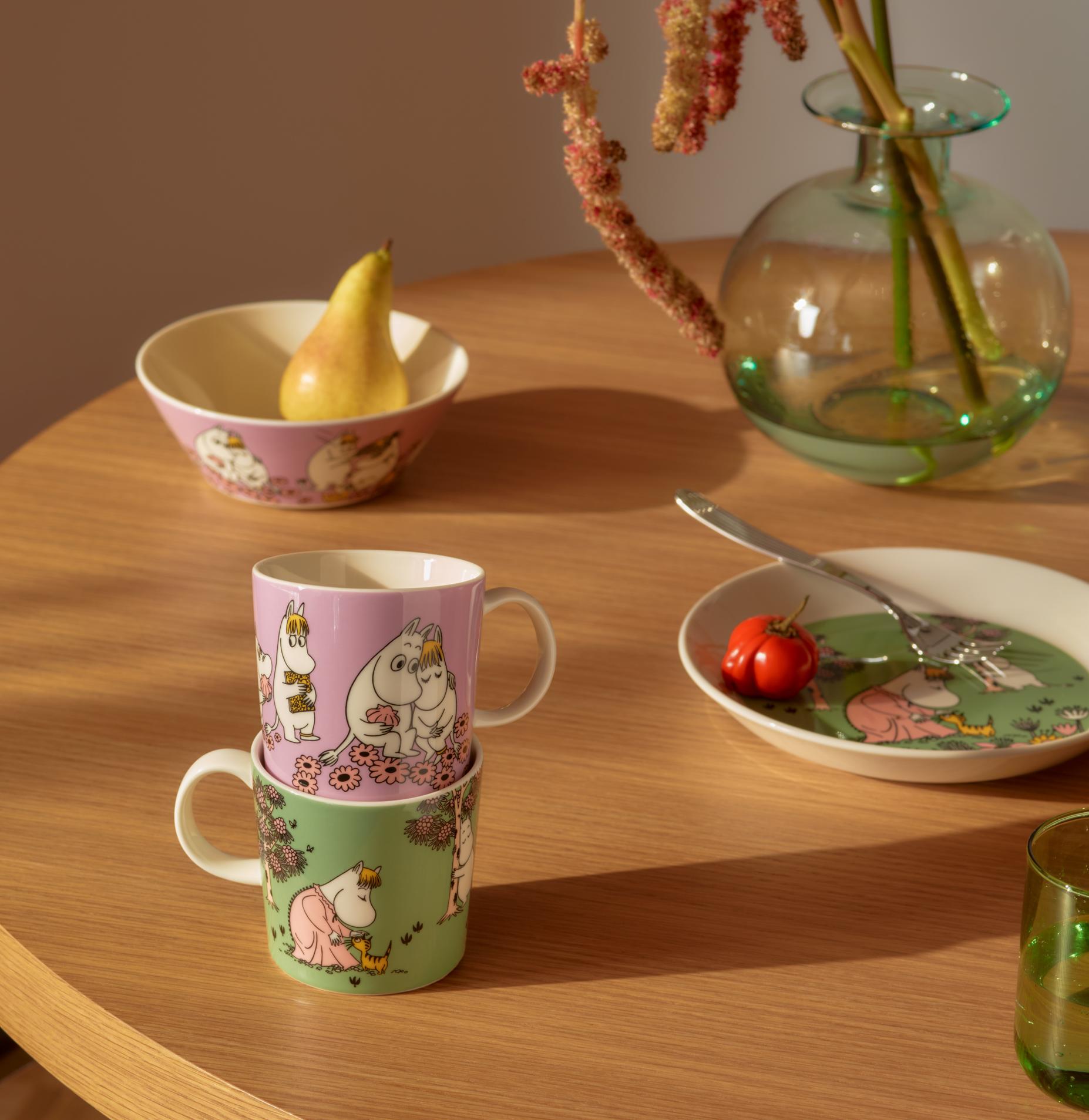 Muminmuggar staplade på ett träbord med en matchande skål, tallrik, päron och rosa blommor.