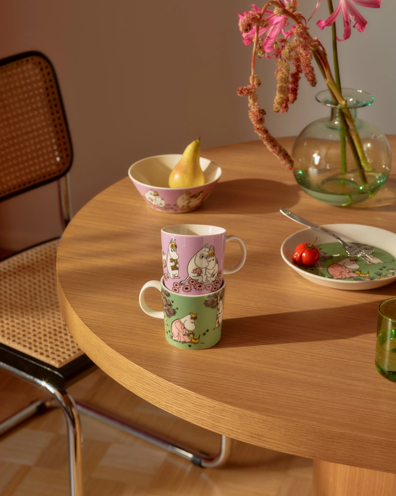 Two stacked Moomin mugs and matching dishes on a wooden table with a pear and flowers.
