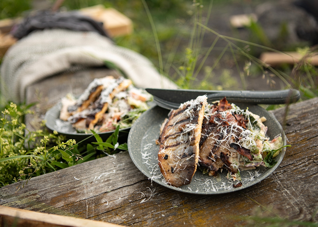Grillad fisk med riven ost och sallad serverad utomhus på en rustik träbänk i naturen.