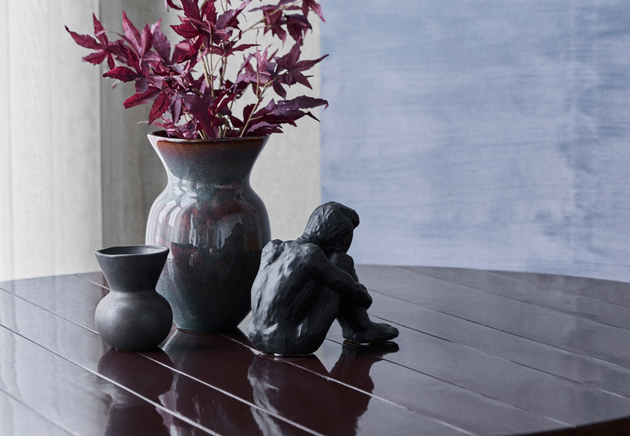 Mørk vase med burgunderrøde blader, liten keramikkvase og svart skulptur på et blankt spisebord.