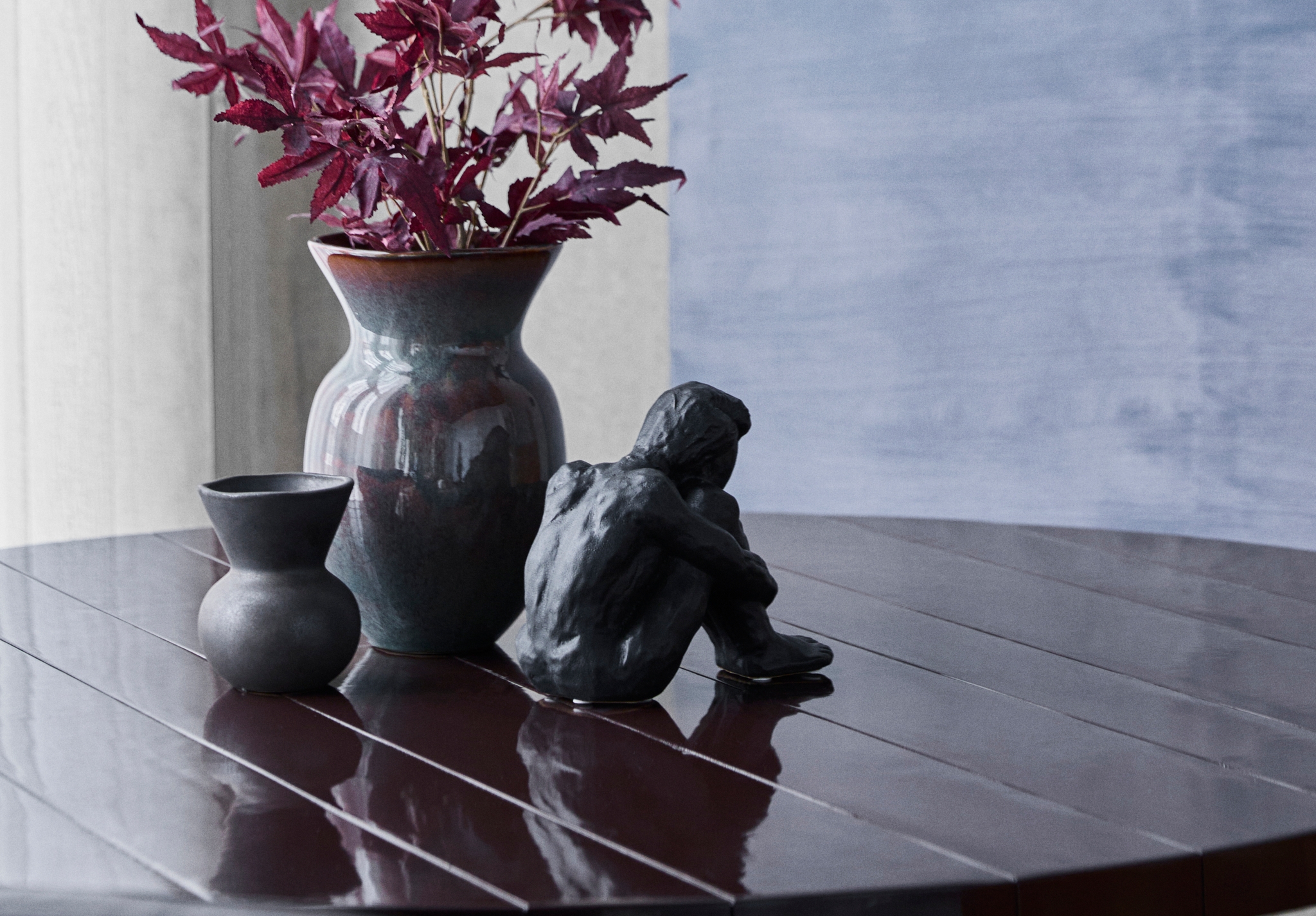Mørk vase med burgunderrøde blader, liten krukke og svart skulptur på et blankt spisebord.