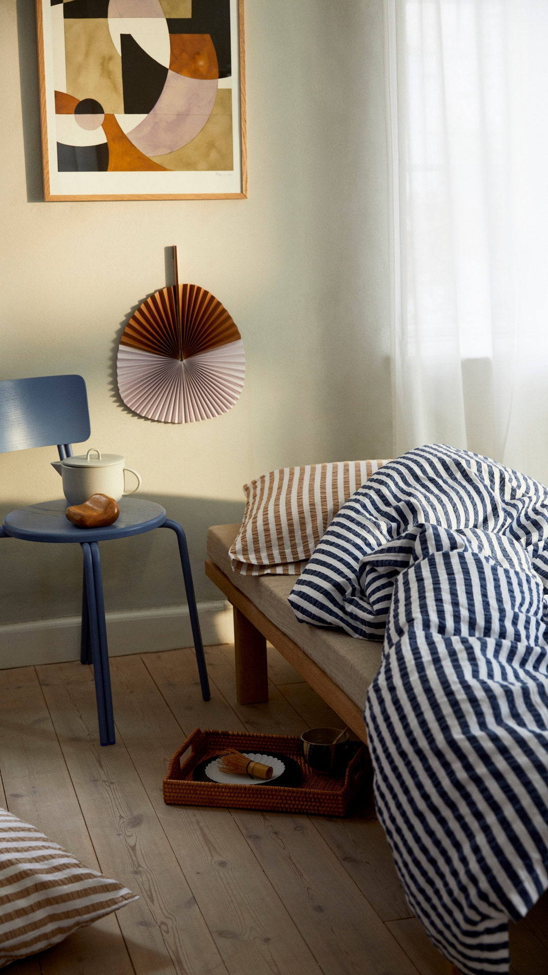 Sovrum med en säng med blått randigt täcke och röd randig kudde, men en blå stol bredvid och röd solfjäder på väggen