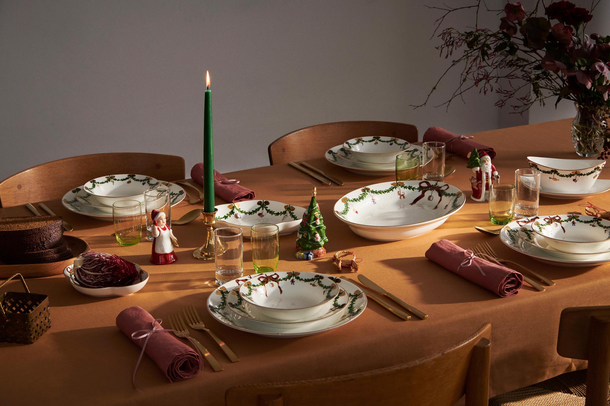 Ett festligt matbord med vita fat prydda med gröna girlanger, ett tänt ljus, julfigurer och en varm orange duk.