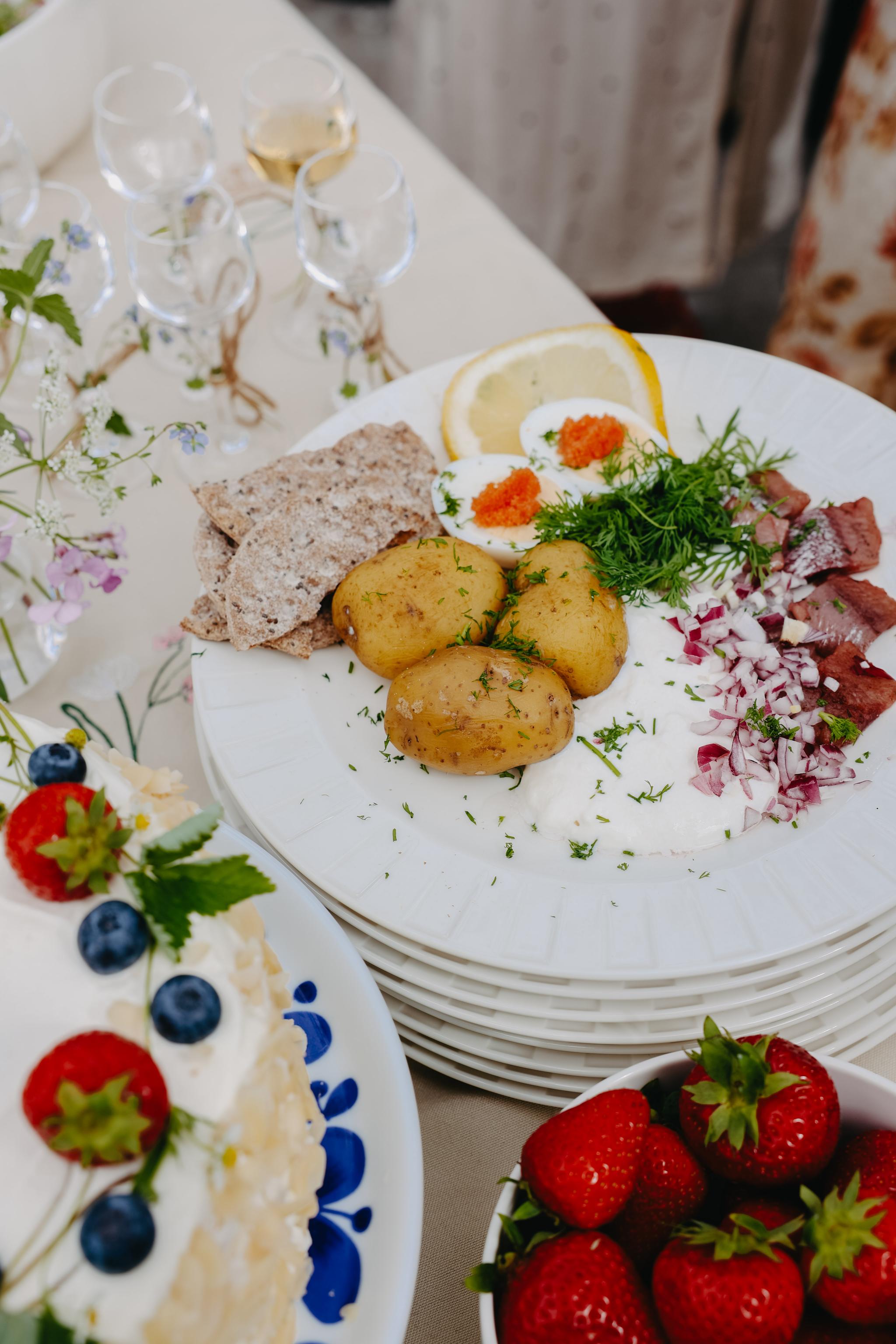 Somrig svensk midsommartallrik med färskpotatis, sill, ägg, knäckebröd, gräddfil och jordgubbstårta.