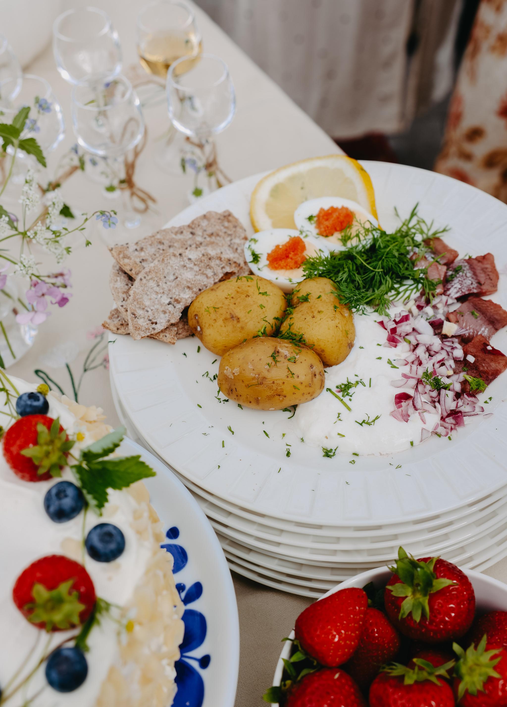 Somrig svensk buffé med sill, färskpotatis, knäckebröd, ägg med rom, jordgubbar och gräddtårta med bär på ett dukat bord.