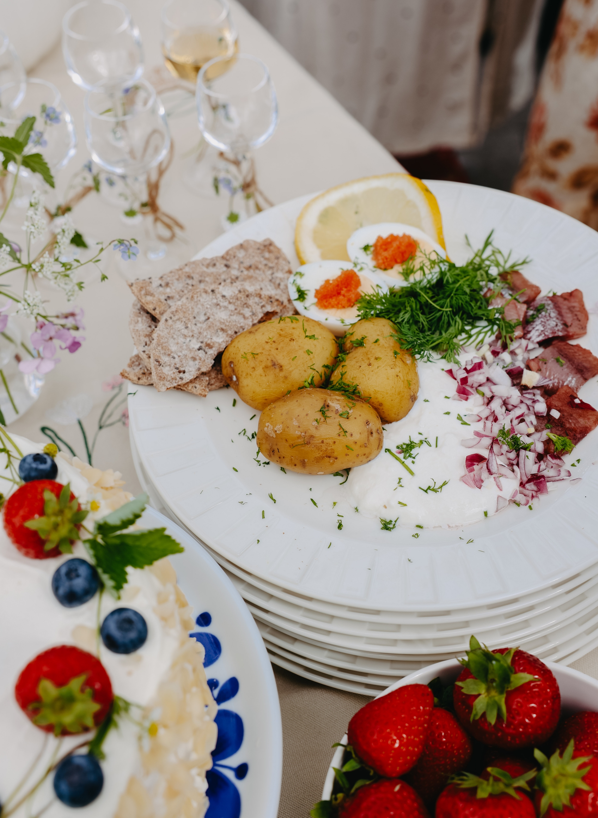 Somrig svensk buffé med sill, färskpotatis, ägg med rom, knäckebröd, jordgubbar och gräddtårta på dukat bord.