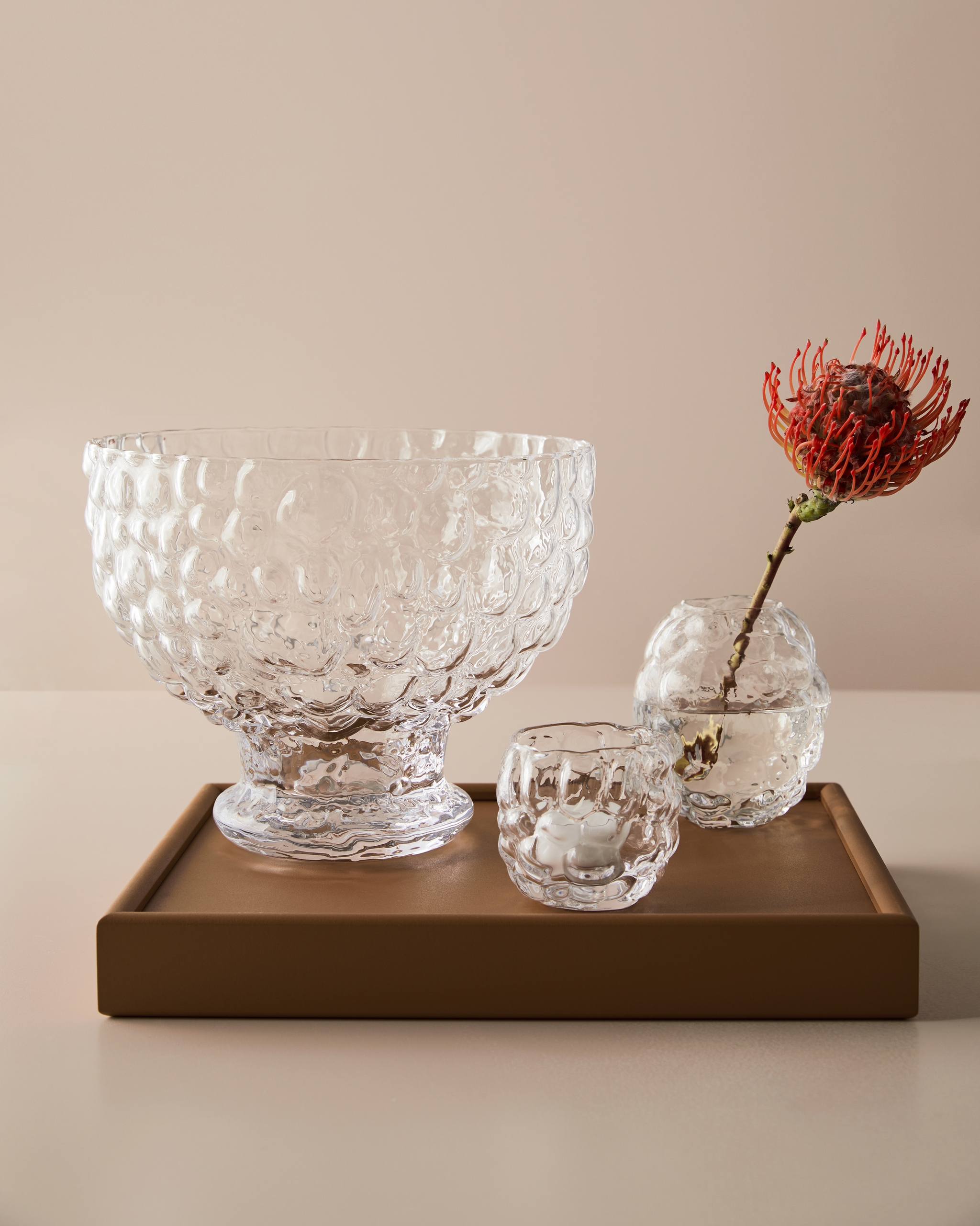 Handgjorda glasskålar och en glasvas med en röd blomma på en brun bricka.