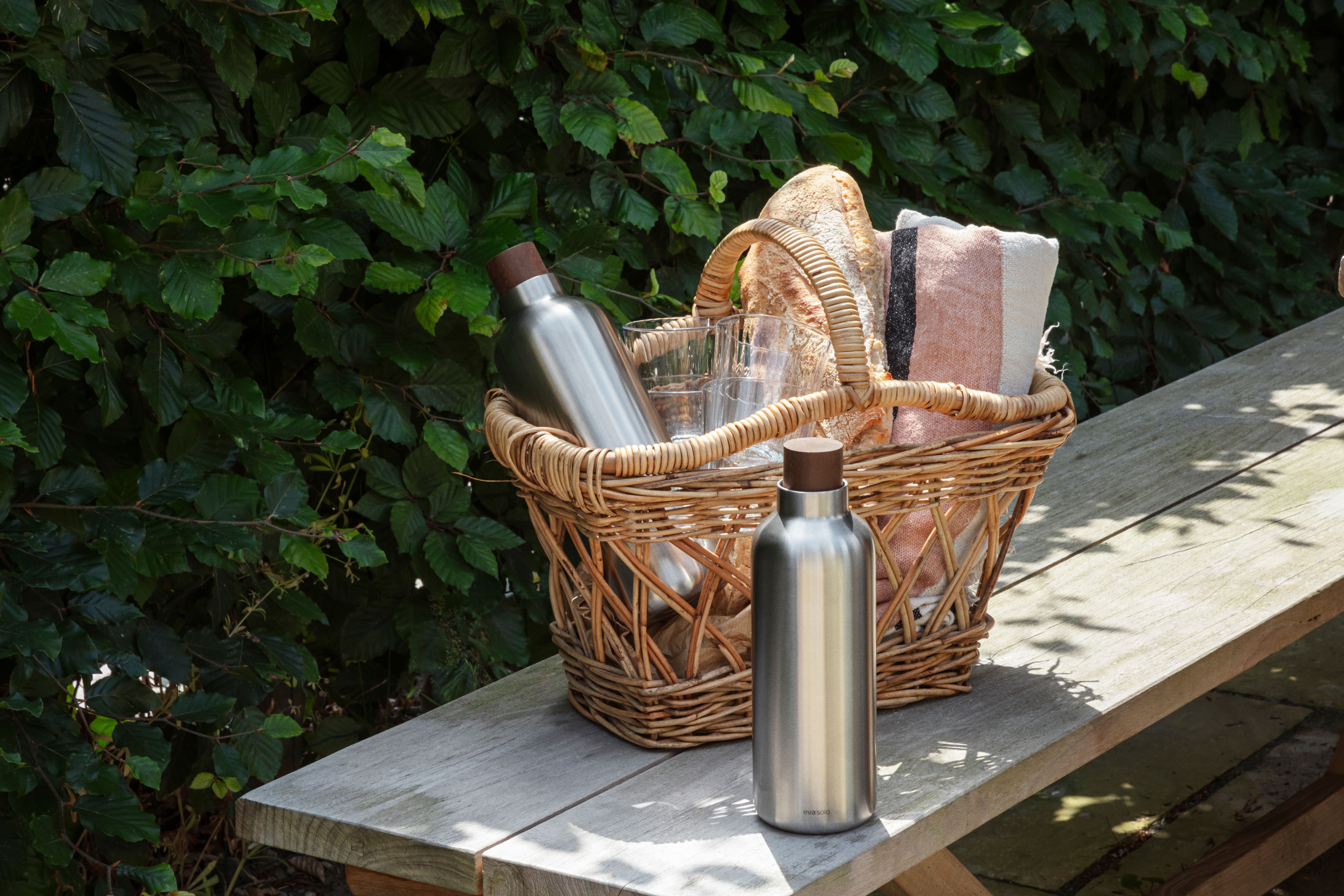 En picknickkorg med bröd, en randig handduk och två rostfria flaskor står på en träbänk framför grönska