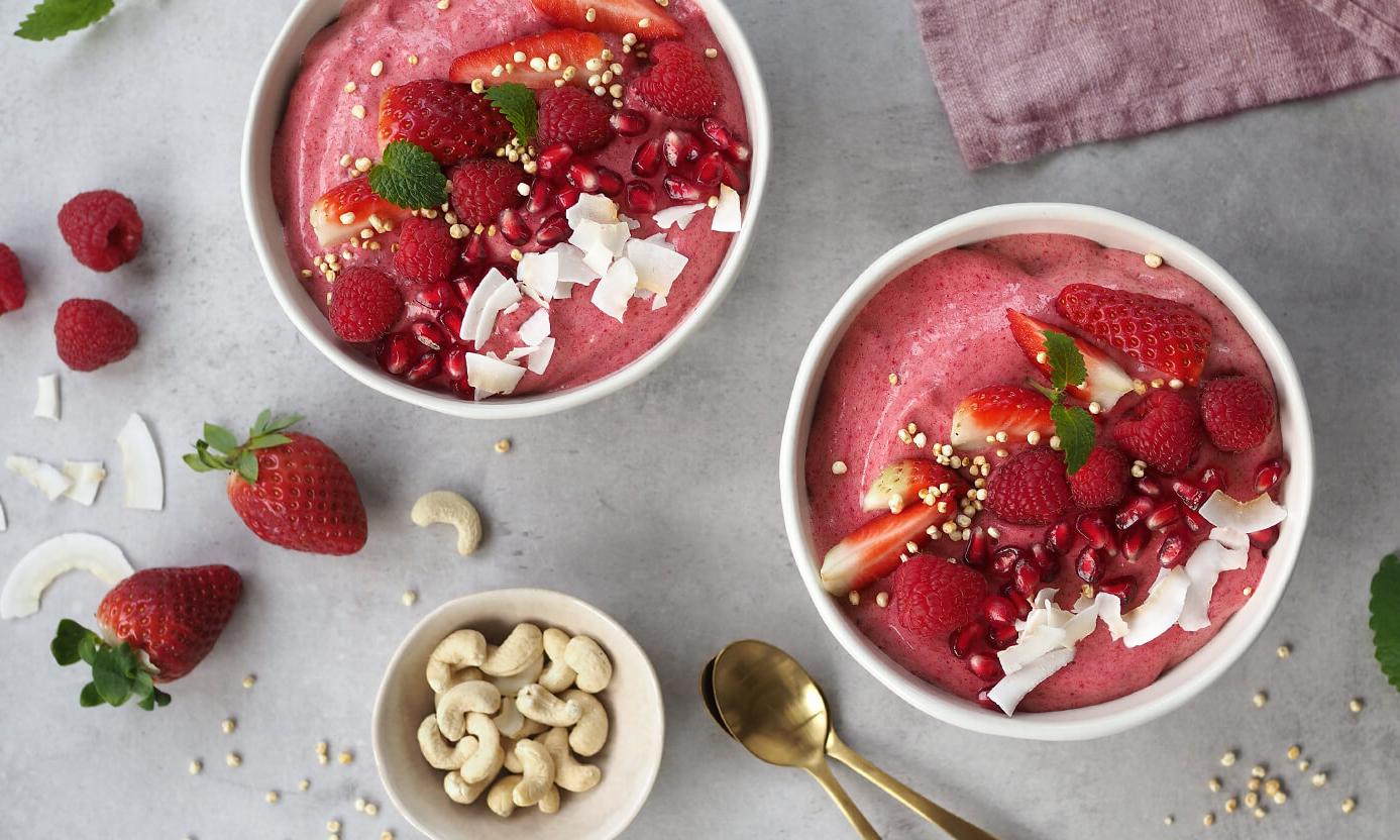 Kaksi vaaleanpunaista smoothie-kulhoa koristeltuna mansikoilla, vadelmilla, granaattiomenansiemenillä ja kookoslastuilla harmaalla taustalla.