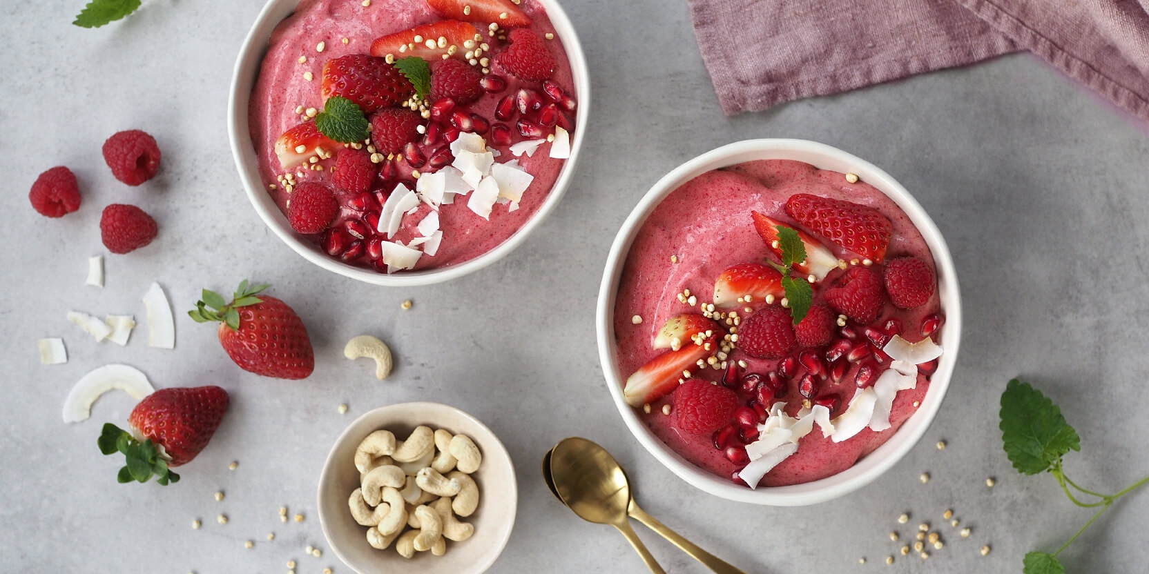 Kaksi vaaleanpunaista smoothie-kulhoa on koristeltu mansikoilla, vadelmilla, granaattiomenansiemenillä ja kookoslastuilla harmaalla taustalla.