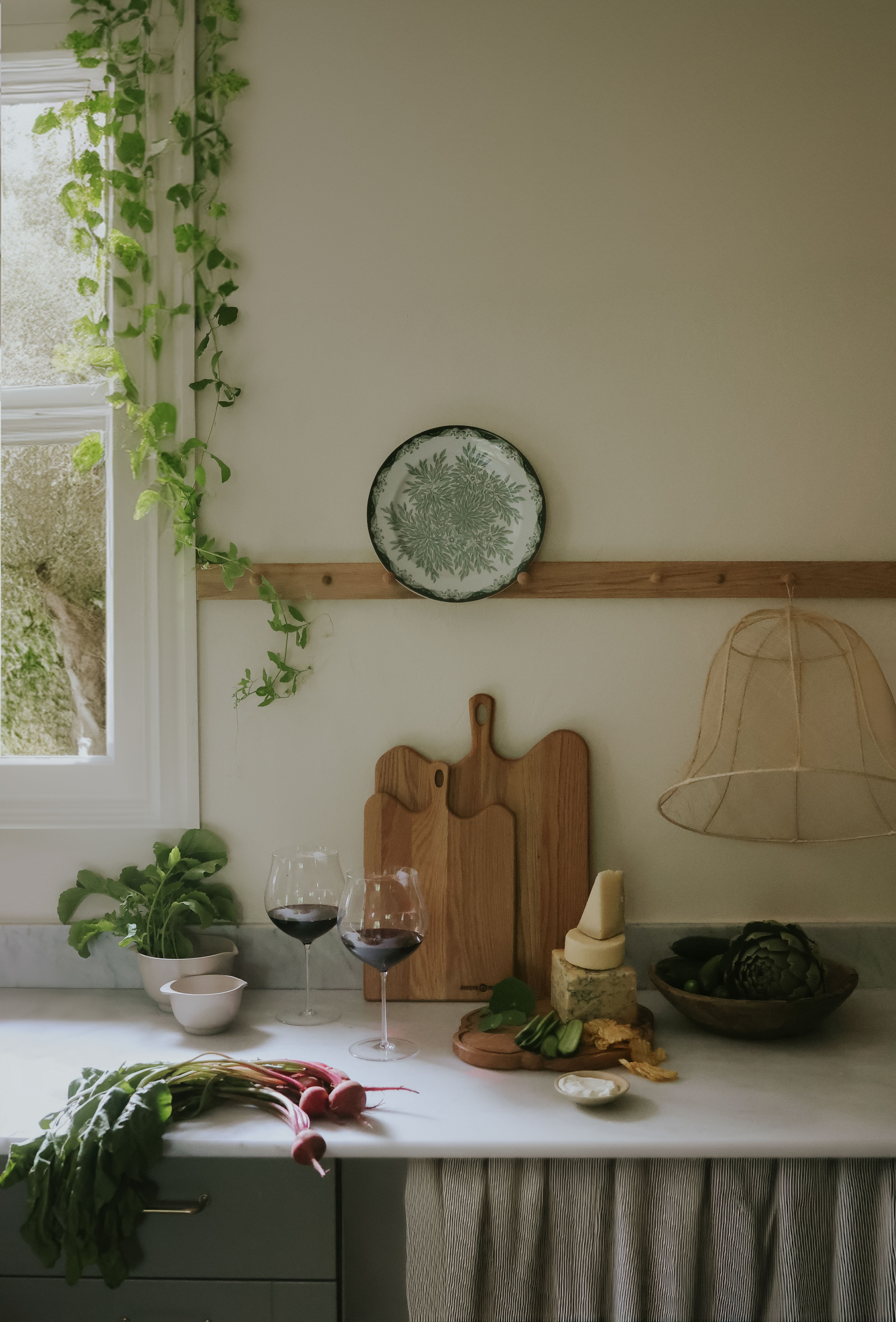 A kitchen counter by a window with trailing green vines, featuring fresh radishes, two wine glasses, a stack of cheeses, artichokes, and wooden cutting boards.