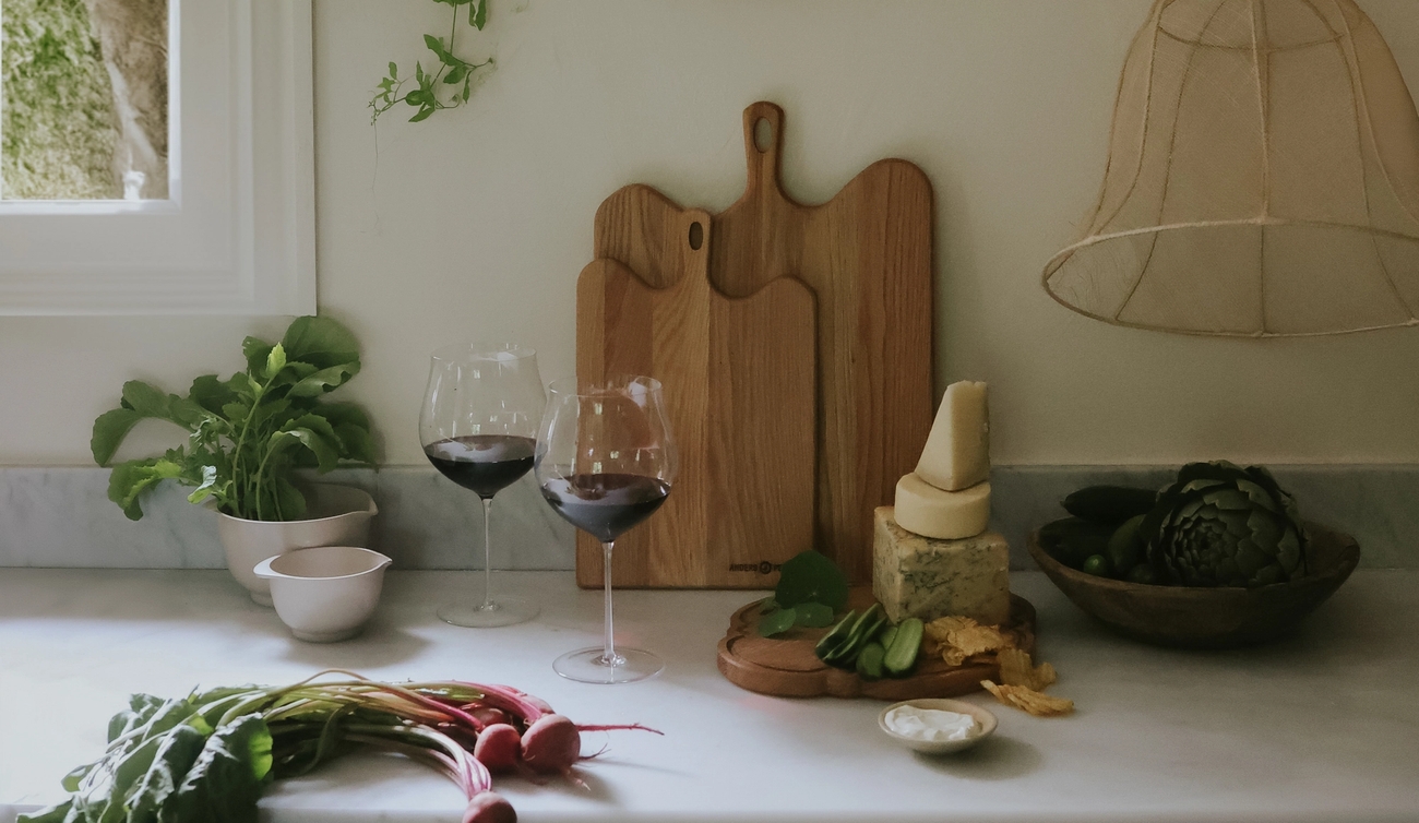 A kitchen counter by a window with trailing green vines, featuring fresh radishes, two wine glasses, a stack of cheeses, artichokes, and wooden cutting boards.