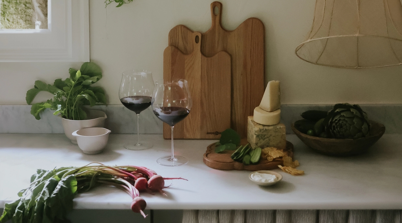 A kitchen counter with red wine, fresh radishes, cheese, and artichokes, with wooden cutting boards and vines by a window.