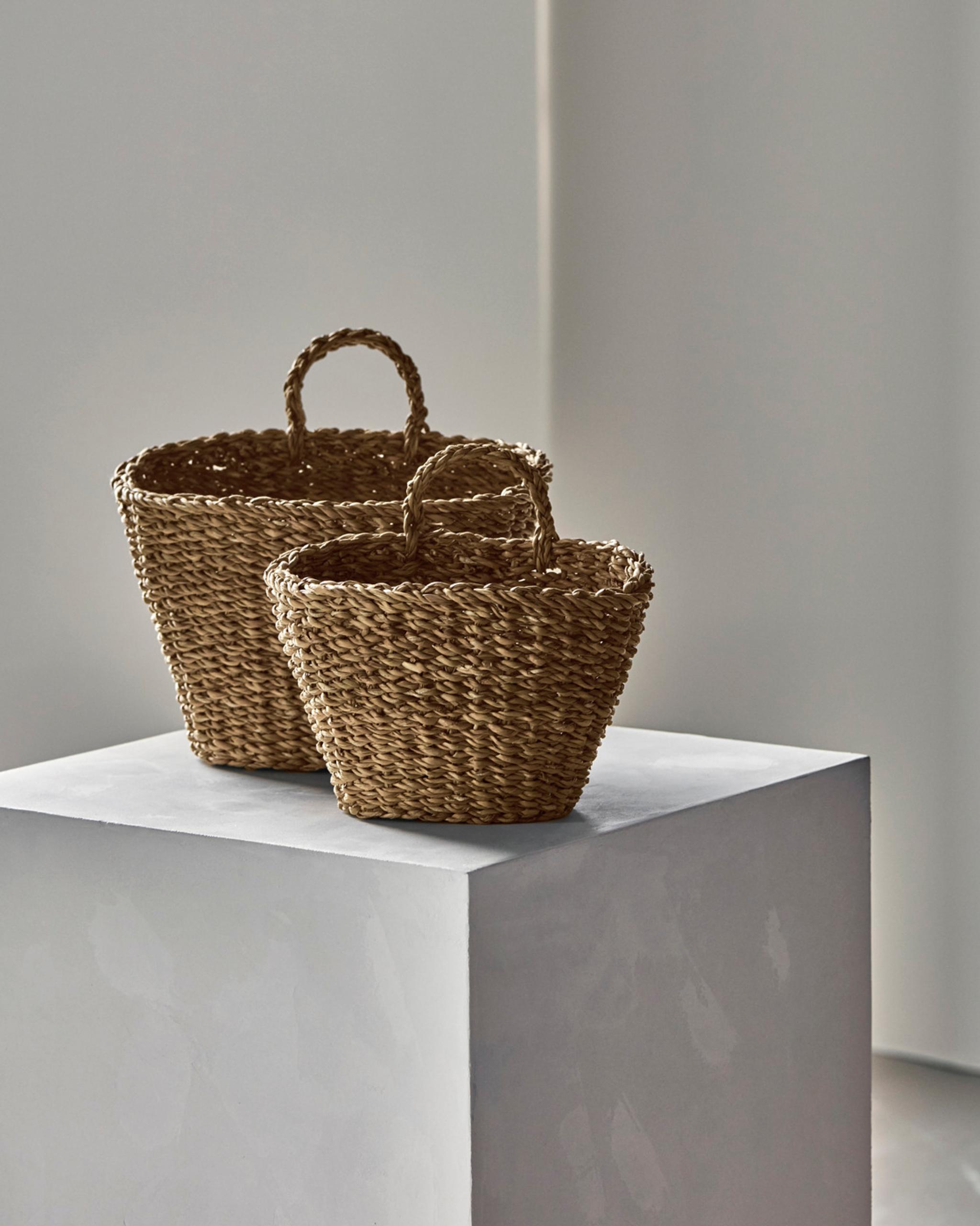 Två flätade korgar i naturmaterial står på ett vitt podium i en minimalistisk inredningsmiljö.