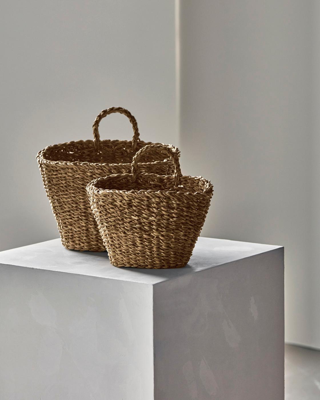 Två flätade korgar i naturmaterial står på ett vitt podium i en minimalistisk inredningsmiljö.