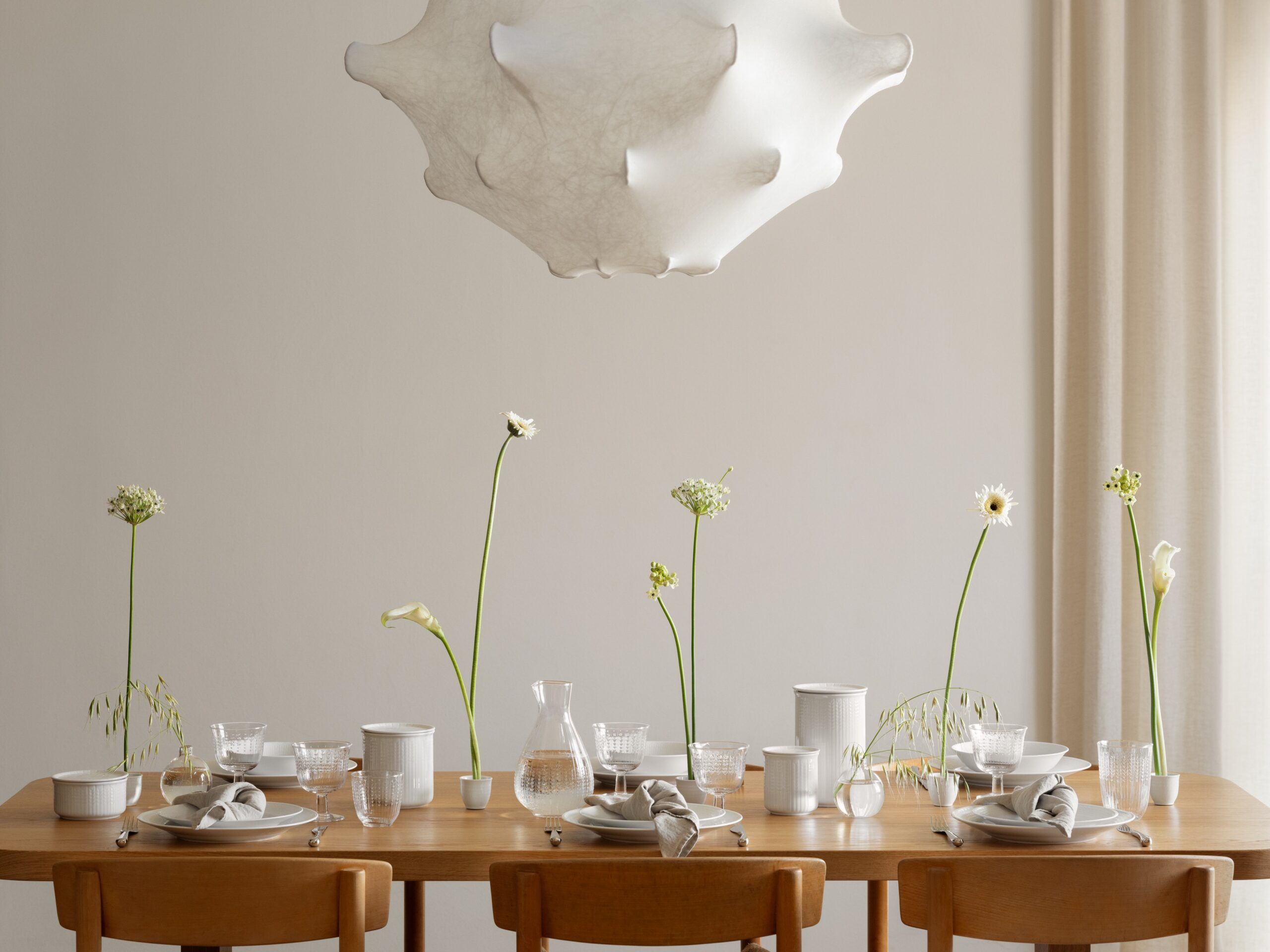 Ett minimalistiskt matbord dukat med vitt porslin, skira vita blommor och en skulptural lamparmatur.