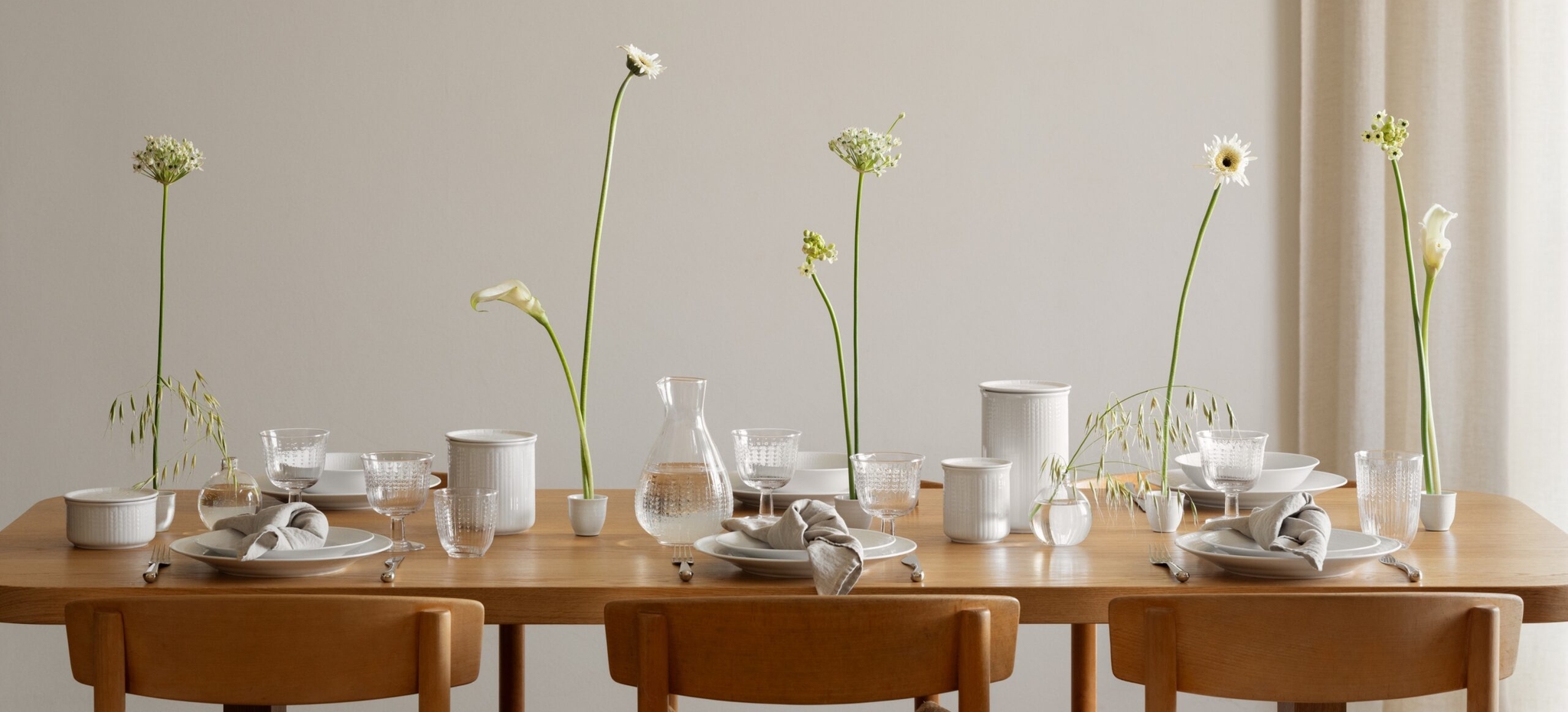 Et spisebord i tre er dekket med hvitt servise, glass og minimalistiske, høye hvite blomster, under en unik, skulpturell hvit taklampe