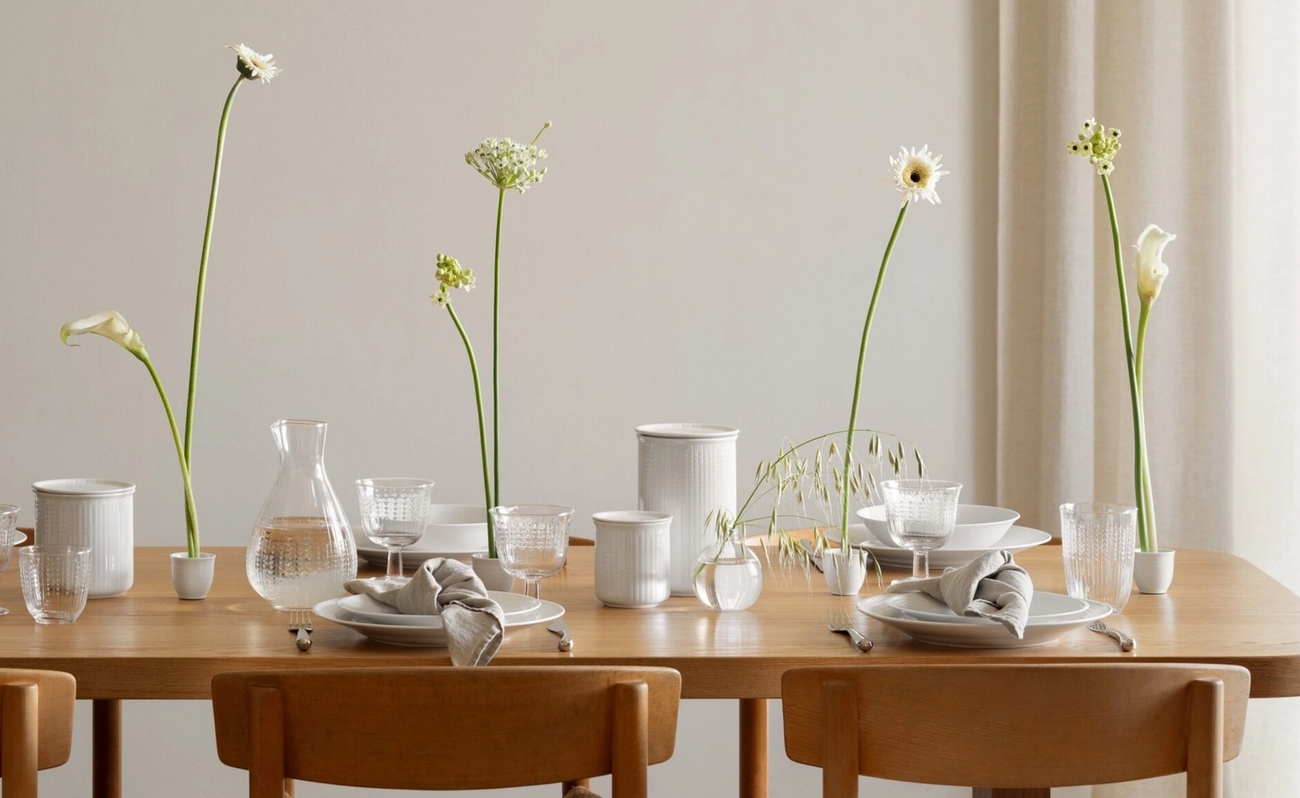 Ett minimalistiskt matbord dukat med vitt porslin, skira vita blommor och en skulptural lamparmatur.