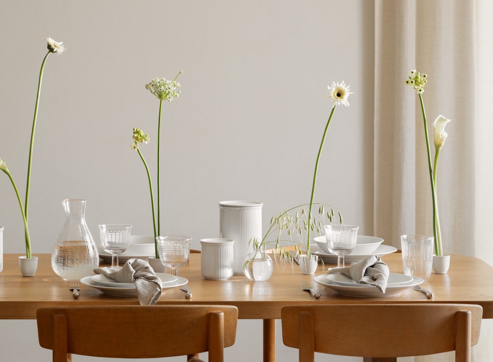 Et spisebord i tre er dekket med hvitt servise, glass og minimalistiske, høye hvite blomster, under en unik, skulpturell hvit taklampe