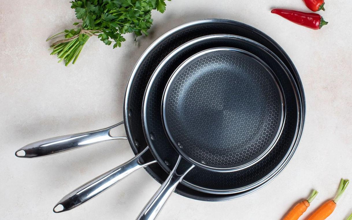 Kolme erikokoista paistinpannua on pinottu keittiötasolle tuoreiden porkkanoiden, chilien ja persiljan viereen.