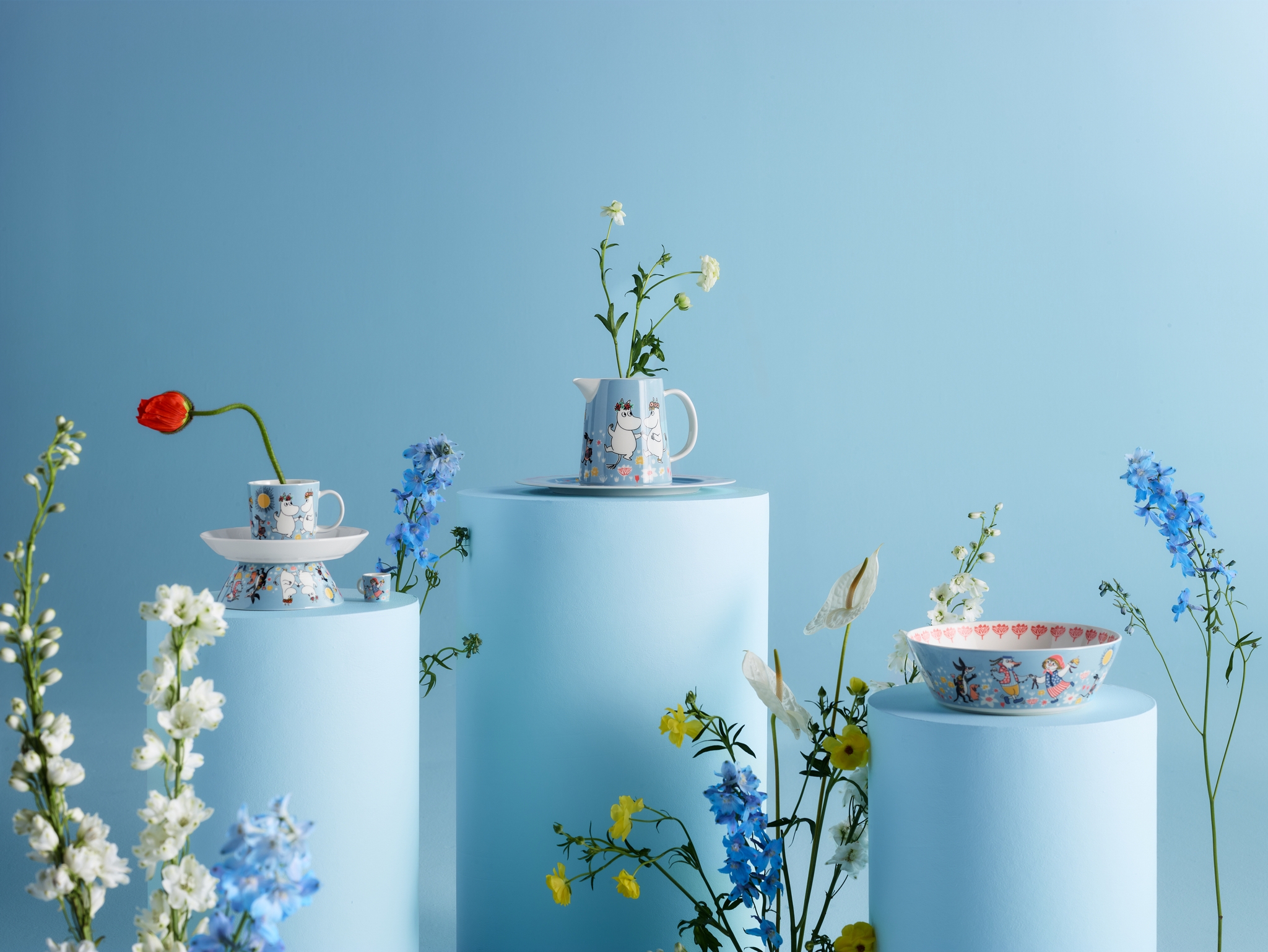 Two stacked Moomin mugs and matching dishes on a wooden table with a pear and flowers.
