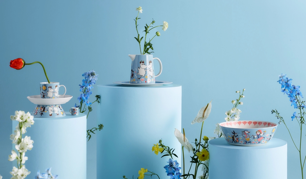 Mumininspirerat porslin med blommor arrangerat på ljusblå piedestaler mot en blå bakgrund.