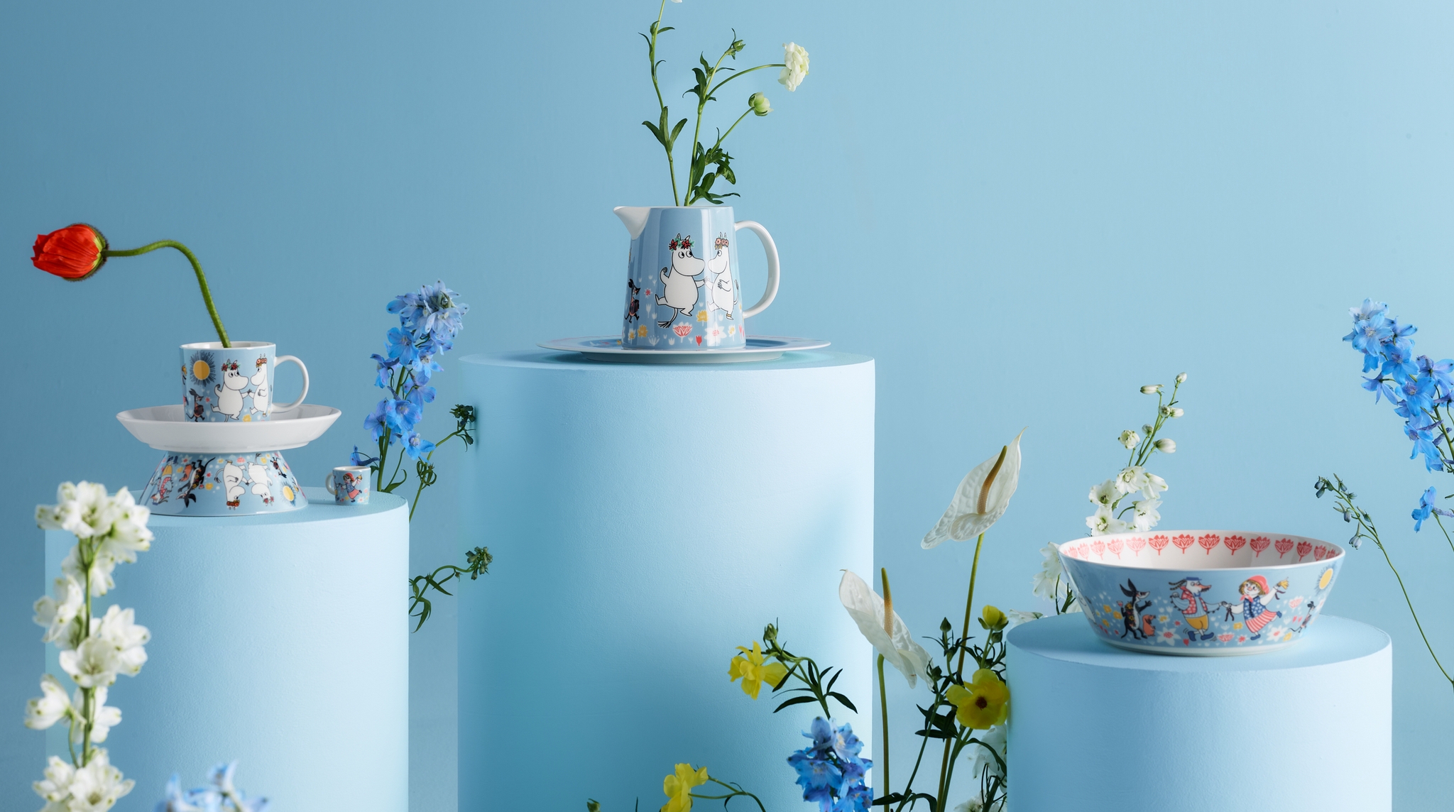 Pastellfärgade Mumin-muggar och skålar med blommor arrangerade på ljusblå podier mot en blå bakgrund.