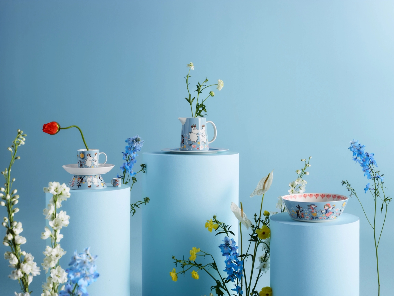 Two stacked Moomin mugs and matching dishes on a wooden table with a pear and flowers.