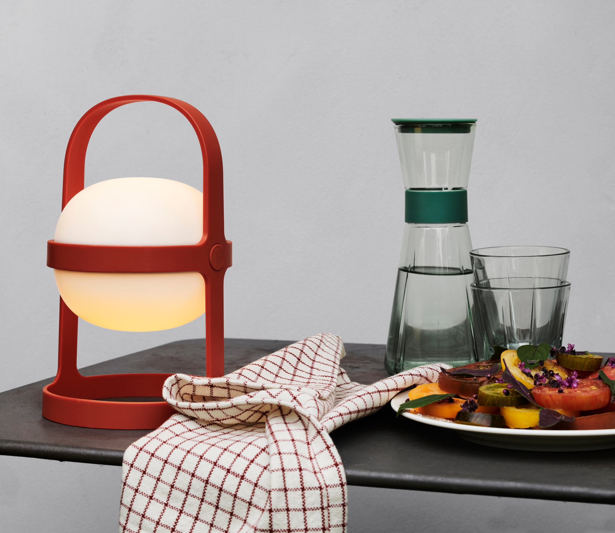 En glødende rød bærbar lyktelampe, en rutete serviett, en glasskaraffel med to glass og en fargerik salat på en mørk overflate mot en lys grå vegg