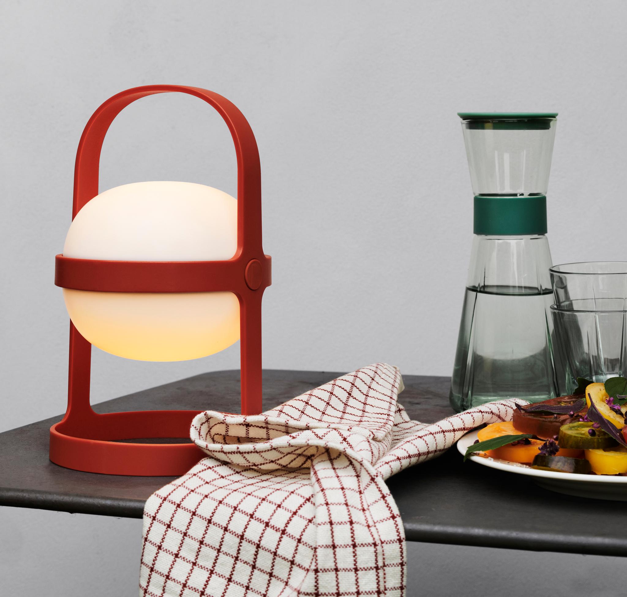 En glødende rød bærbar lyktelampe, en rutete serviett, en glasskaraffel med to glass og en fargerik salat på en mørk overflate mot en lys grå vegg