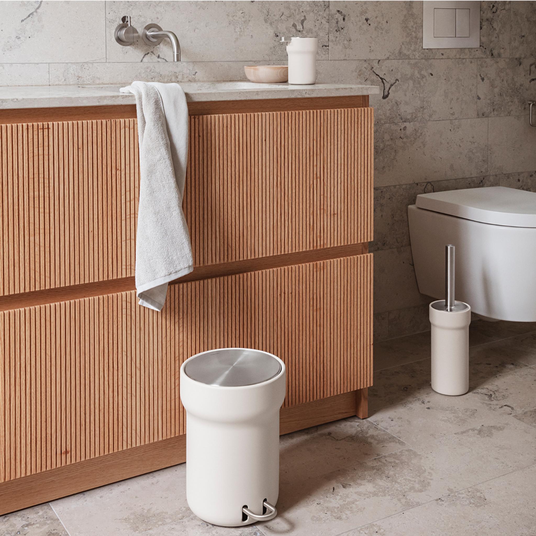 Modernt badrum med räfflat badrumsskåp i trä, vit handduk, krämfärgad papperskorg, toalettborste och vägghängd toalett.