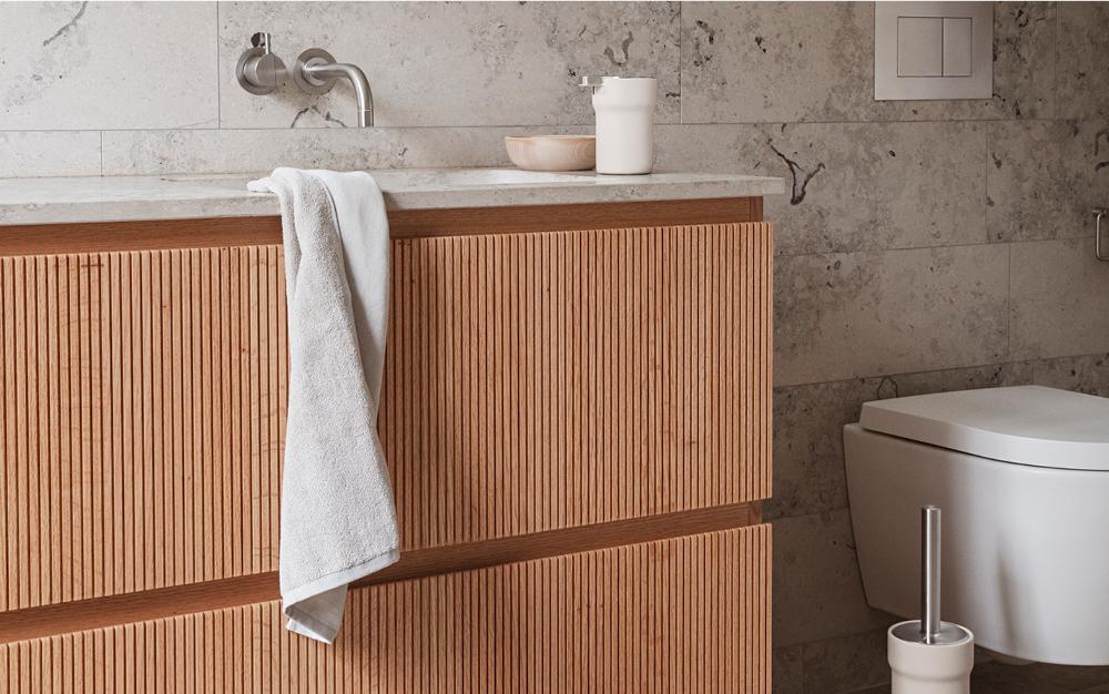 Modernt badrum med ett räfflat badrumsskåp i ljust trä, vit bänkskiva, vit pedalhink och toalettborste.