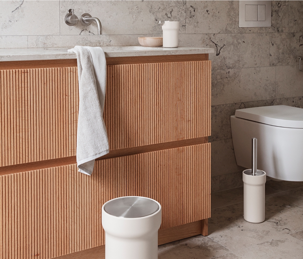 Modernt badrum med ett räfflat badrumsskåp i ljust trä, vit bänkskiva, vit pedalhink och toalettborste.